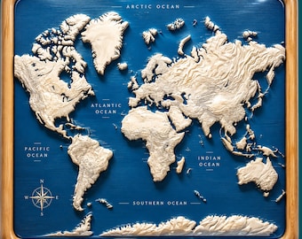 May include: A square wall map featuring raised, cream-colored topographical landmasses set against a deep blue ocean background. The map is encased in a smooth, light-toned wooden frame and includes labels for the Arctic, Atlantic, Pacific, Indian, and Southern Oceans, along with a compass rose in the bottom left corner.