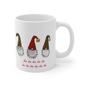 May include: White ceramic mug featuring three Christmas gnomes with red, green, and gold accents. Each gnome has a long white beard and a pointed hat. The text "La-la-la-la-la" is printed in red below the gnomes.