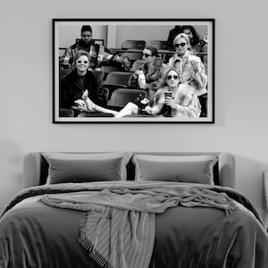 May include: Black and white framed print of a group of people wearing sunglasses, seated in a stadium. The image is displayed above a bed with gray bedding and a gray throw blanket.