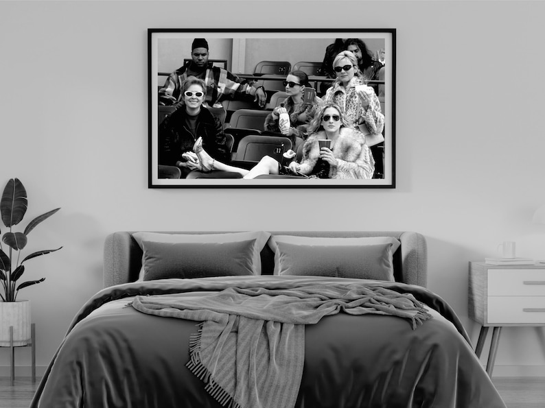 May include: Black and white framed print of a group of people wearing sunglasses, seated in a stadium. The image is displayed above a bed with gray bedding and a gray throw blanket.