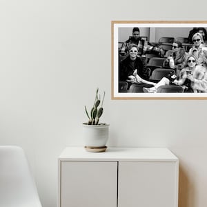May include: Black and white photograph of a group of people sitting in stadium seats. The people are wearing sunglasses and coats. The photograph is framed in a light-colored wooden frame.
