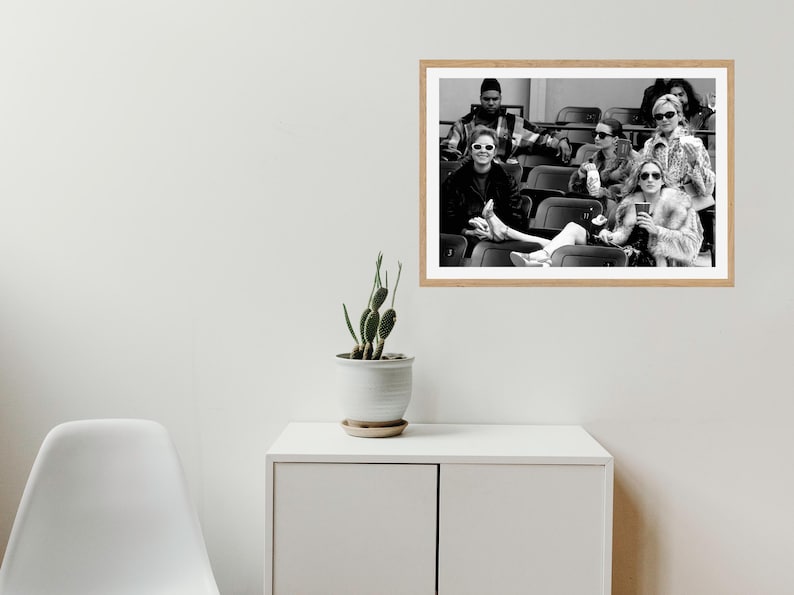 May include: Black and white photograph of a group of people sitting in stadium seats. The people are wearing sunglasses and coats. The photograph is framed in a light-colored wooden frame.