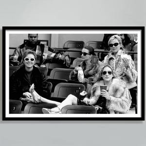 May include: Black and white framed photograph of a group of people in a stadium setting. The subjects are wearing sunglasses and fur coats. The image is a candid shot, capturing a moment in time.