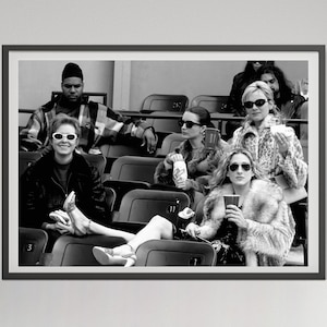 May include: Black and white framed photograph of a group of people seated in stadium seating. Several people are wearing sunglasses and fur coats. One person is holding a cup. The image is a candid shot.