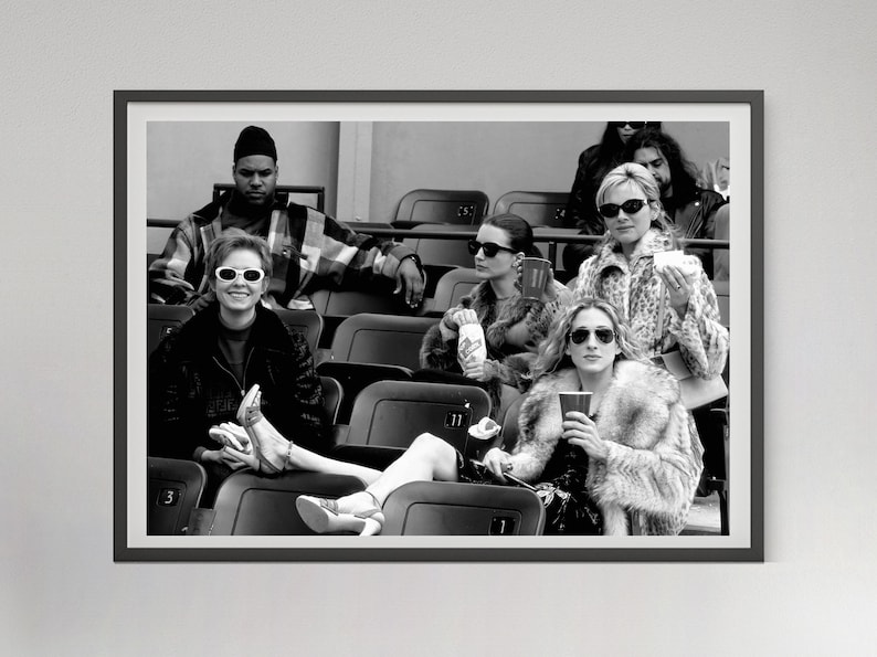 May include: Black and white framed photograph of a group of people seated in stadium seating. Several people are wearing sunglasses and fur coats. One person is holding a cup. The image is a candid shot.