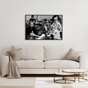 May include: Black and white framed photograph of a group of people wearing sunglasses, seated in stadium seating. The photograph is hanging above a beige sofa with pillows and a blanket. Two round coffee tables are in front of the sofa.
