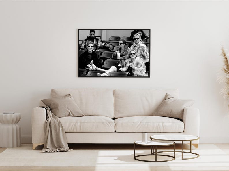 May include: Black and white framed photograph of a group of people wearing sunglasses, seated in stadium seating. The photograph is hanging above a beige sofa with pillows and a blanket. Two round coffee tables are in front of the sofa.