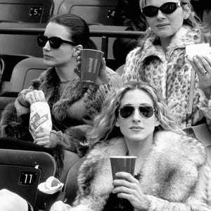 May include: Black and white image of three people wearing sunglasses and fur coats, seated in stadium seating. One person holds a bag of popcorn, another holds a red cup, and the third holds a small white container. The image is in black and white.