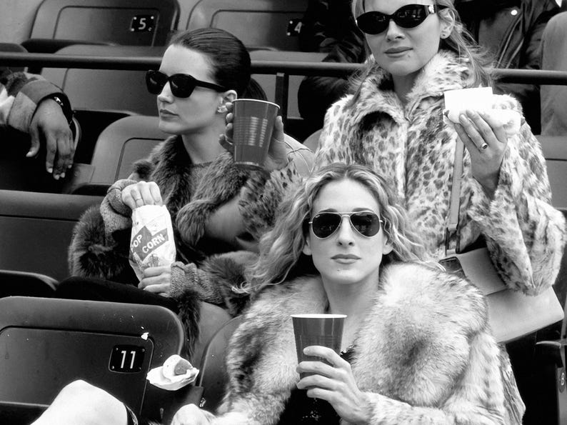 May include: Black and white image of three people wearing sunglasses and fur coats, seated in stadium seating. One person holds a bag of popcorn, another holds a red cup, and the third holds a small white container. The image is in black and white.