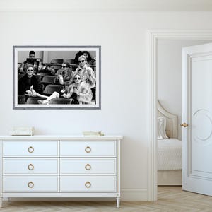 May include: Black and white framed photograph of a group of people wearing sunglasses, seated in stadium seating. The photograph is hanging above a white dresser with six drawers. A partially open white door reveals a bedroom.