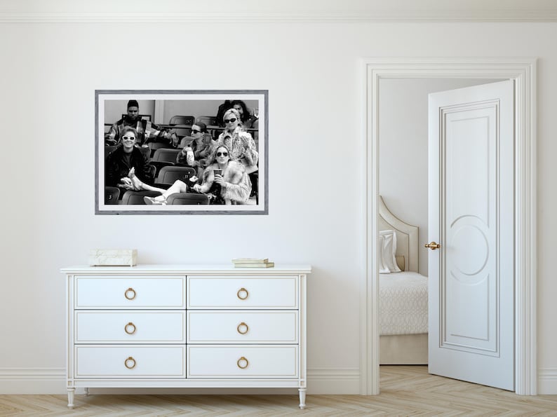 May include: Black and white framed photograph of a group of people wearing sunglasses, seated in stadium seating. The photograph is hanging above a white dresser with six drawers. A partially open white door reveals a bedroom.