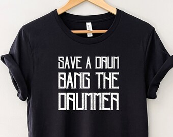 Puede incluir: Camiseta negra con el texto blanco "SAVE A DRUM BANG THE DRUMMER" en una fuente en negrita y en bloques. La camiseta está colgada de una percha de madera sobre un fondo blanco liso. Las mangas están dobladas.