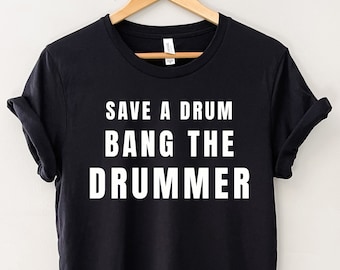 Puede incluir: Camiseta negra con el texto blanco "SAVE A DRUM BANG THE DRUMMER". La camiseta está colgada en una percha de madera. Las mangas están dobladas.