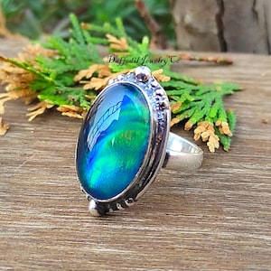 May include: Silver ring featuring an oval, iridescent gemstone in blue and green hues. The ring has a detailed setting and a smooth band. The text "Daffodil Jewelry Co" is visible. The ring is a piece of jewellery.