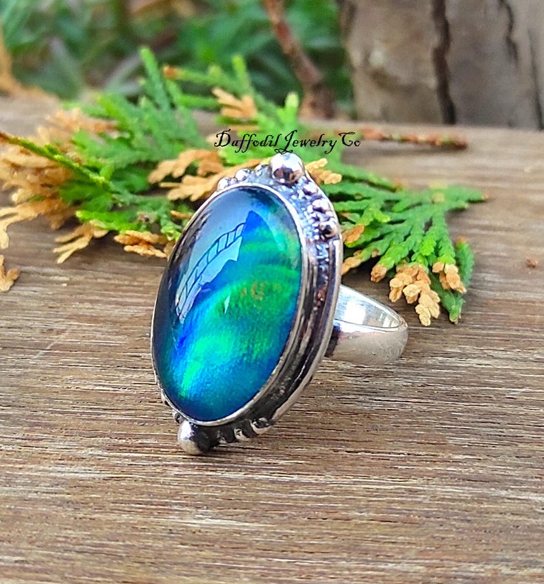 May include: Silver ring featuring an oval, iridescent gemstone in blue and green hues. The ring has a detailed setting and a smooth band. The text "Daffodil Jewelry Co" is visible. The ring is a piece of jewellery.