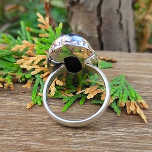 May include: A silver ring featuring a large, oval, black gemstone. The ring's band is smooth and polished. The gemstone is set in a textured silver setting. The text "Daffodil Jewelry Co" is visible on the ring.