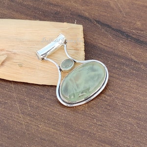 May include: A silver pendant with a light green oval gemstone and a smaller round gemstone. The pendant has a bail for attaching to a chain.