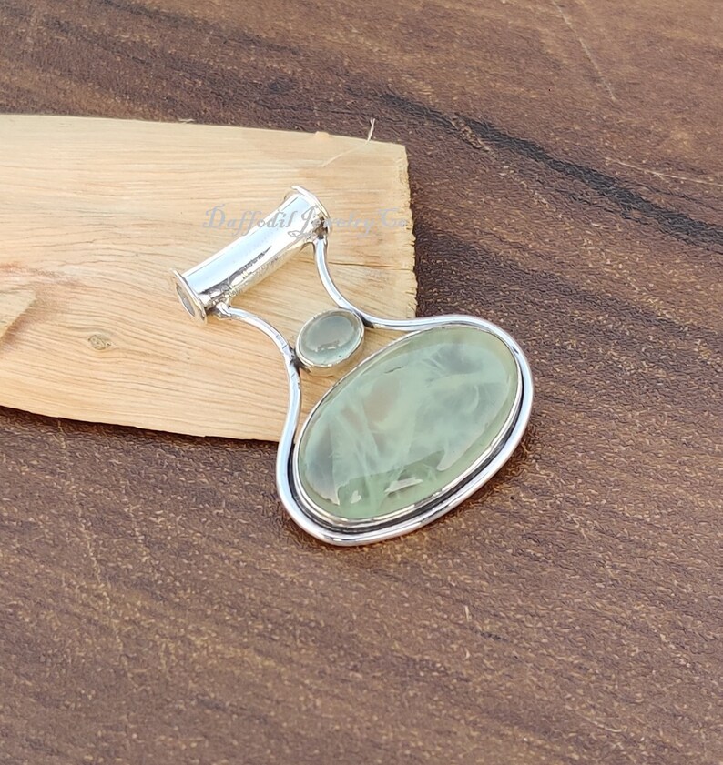 May include: A silver pendant with a light green oval gemstone and a smaller round gemstone. The pendant has a bail for attaching to a chain.