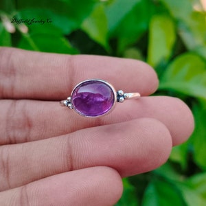 May include: A silver ring featuring a large, oval-shaped, deep purple amethyst gemstone. The ring has a simple band with small decorative silver accents on either side of the gemstone. The ring is held in a hand against a blurred green background.