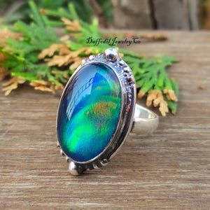 May include: A silver ring featuring an oval iridescent gemstone. The stone has a mix of blue, green, and gold colours. The ring has a detailed, textured setting. The text "Daffodil Jewelry Co" is visible.