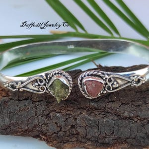 Op de afbeelding: Een zilveren armband met een groene en roze stenen accent. De armband heeft een delicaat bloemmotief.