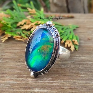 May include: Oval ring featuring an iridescent gemstone in shades of blue, green, and gold, set in a silver-coloured metal band with decorative detailing. The ring is on a wooden surface, with the text "Daffodil Jewelry Co."