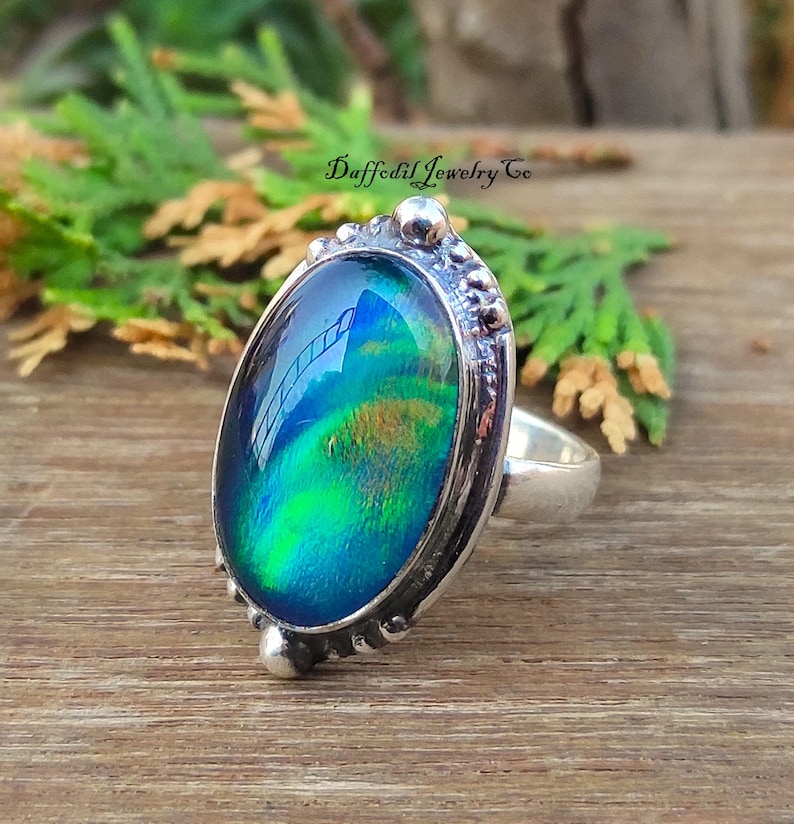May include: Oval ring featuring an iridescent gemstone in shades of blue, green, and gold, set in a silver-coloured metal band with decorative detailing. The ring is on a wooden surface, with the text "Daffodil Jewelry Co."