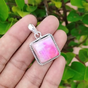 May include: A square pendant featuring a pink and white stone, set within a silver-coloured metal frame. The frame has a twisted rope design. The pendant includes a small loop at the top for a chain.