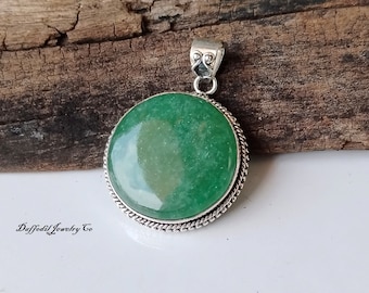 May include: A round green gemstone pendant with a silver bezel setting. The pendant is on a wooden surface.