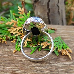 May include: A silver ring featuring a large, oval, black gemstone. The ring's band is smooth and polished. The gemstone is set in a textured, silver bezel. The text "Daffodil Jewelry Co" is visible on the ring.