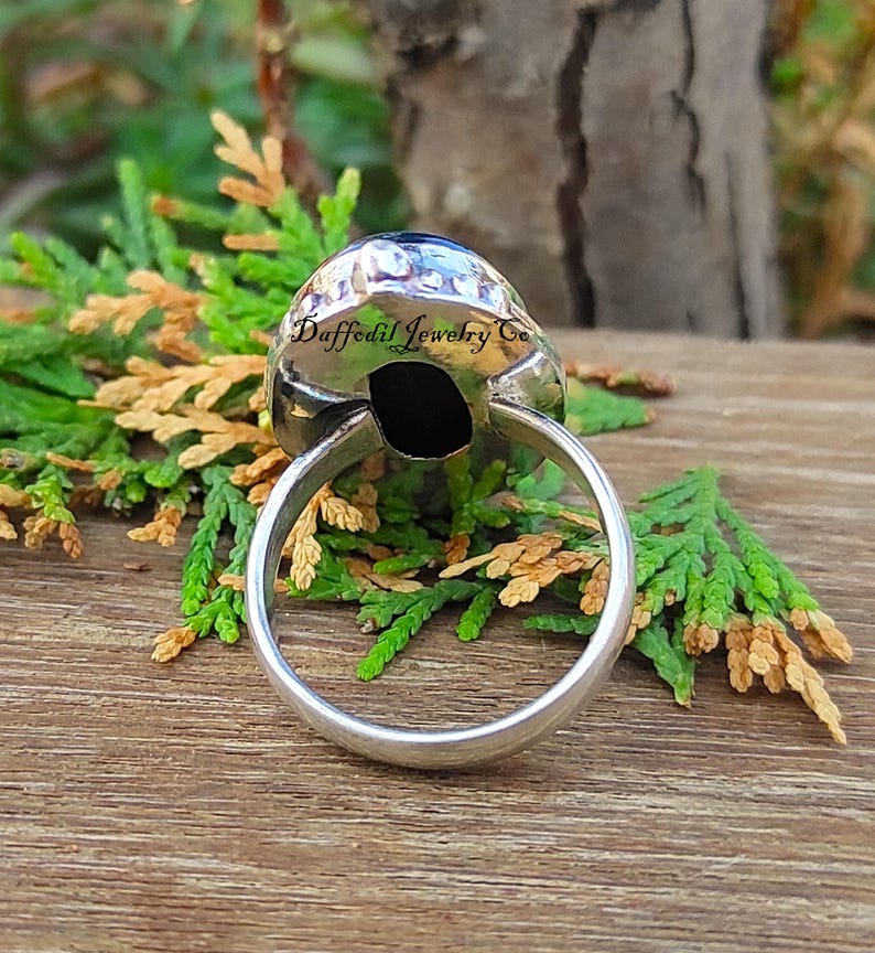 May include: A silver ring featuring a large, oval, black gemstone. The ring's band is smooth and polished. The gemstone is set in a textured, silver bezel. The text "Daffodil Jewelry Co" is visible on the ring.