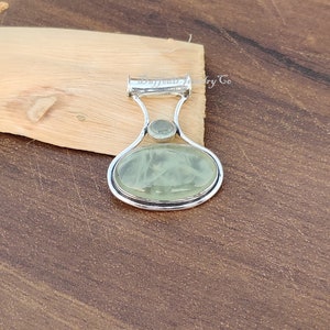 May include: A silver pendant with a light green oval-shaped gemstone and a smaller round gemstone. The pendant has a bail for attaching to a chain.