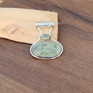 May include: A silver pendant with a light green oval-shaped gemstone and a smaller round gemstone. The pendant has a bail for attaching to a chain.