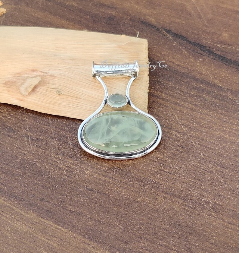 May include: A silver pendant with a light green oval-shaped gemstone and a smaller round gemstone. The pendant has a bail for attaching to a chain.