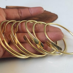 May include: A set of five gold-toned metal bangles, stacked and overlapping on a hand. The bangles are simple and smooth, with a slightly rounded shape.