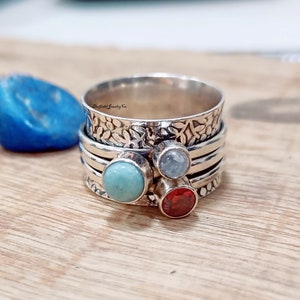 May include: A silver spinner ring with three colorful gemstones: turquoise, light blue, and red. The top band features a floral design. The ring is displayed on a wooden surface.