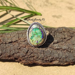 May include: A silver ring with a green and purple opal gemstone set in a twisted silver band. The ring is on a piece of wood with the text "Daffodil Jewelry Co" in the background.