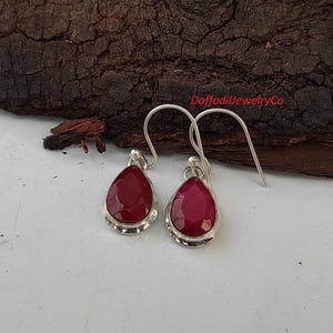 May include: A pair of silver dangle earrings featuring red teardrop-shaped gemstones. The earrings have a simple design and are ideal for everyday wear.