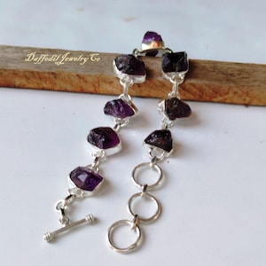 May include: A silver chain bracelet with a toggle clasp features six purple amethyst gemstones. The amethyst stones are rough and irregular in shape, giving the bracelet a natural and rustic look.