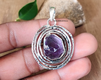 May include: A silver pendant with a purple amethyst gemstone set in a circular design with concentric circles. The pendant is made of sterling silver and features a raw, natural amethyst stone.