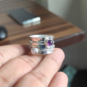 Op de afbeelding: Een zilveren ring met een gehammerde afwerking en drie stenen. De stenen zijn een paarse amethist, een witte parel en een zwarte hartvormige steen.