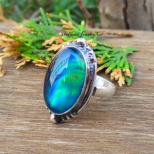 May include: Silver ring featuring an oval iridescent gemstone in blue and green hues. The ring has a detailed setting and a smooth band. The text "Daffodil Jewelry Co" is visible. The ring is a piece of jewellery.