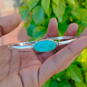 May include: A silver bangle bracelet with a turquoise oval stone set in the center. The bracelet is open and has a simple design.