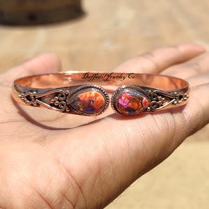 Può includere: Bracciale a polsino in rame con due pietre ovali multicolori. Le pietre presentano sfumature di arancione, rosa e blu. Il bracciale ha un design aperto con elementi decorativi in metallo e il testo "Daffodil Jewelry Co".