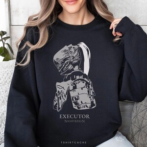 May include: Black sweatshirt with a gray graphic of a helmet and armor. The text "EXECUTOR NIGHTREIGN" is printed below the graphic. The brand name "TSHIRTCACHE" is printed at the bottom of the sweatshirt.