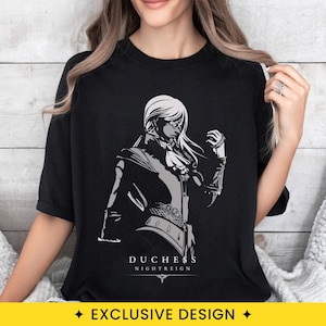 May include: A black short-sleeved t-shirt featuring a graphic print of a character with long hair and an eye covering. The design is rendered in shades of gray and white, with the text DUCHESS NIGHTREIGN printed in white below the illustration.