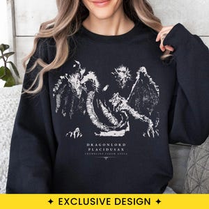 May include: A black crewneck sweatshirt featuring a white graphic print of a multi-headed dragon creature. Below the illustration, the text reads DRAGONLORD PLACIDUSAX CRUMBLING FARUM AZULA with a small decorative symbol underneath the text.