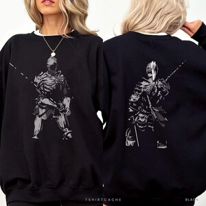 May include: A black long-sleeve sweatshirt featuring a graphic print of a knight in armor on the front and back. The knight is depicted in shades of gray and white, holding a long spear-like weapon. The text TSHIRTSCACHE is printed at the bottom.
