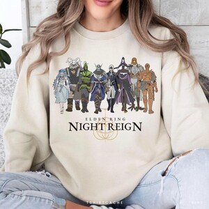 May include: A cream-colored sweatshirt featuring a graphic of characters from Elden Ring. The design includes the text "ELDEN RING NIGHT REIGN" in a bold font, with a golden emblem below the text. The characters are in various colors.