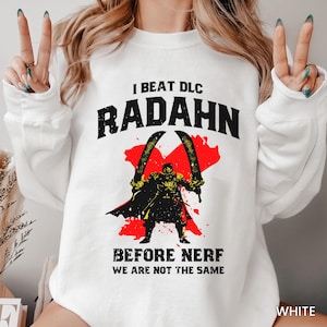 May include: White sweatshirt with a graphic print of a character from the video game Elden Ring. The text on the shirt reads "I beat DLC Radahn before nerf we are not the same".